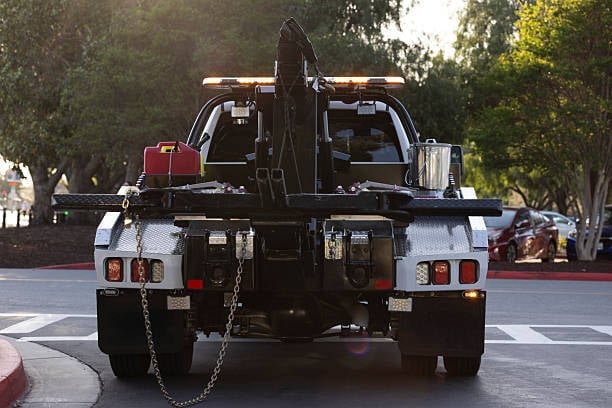 Black tow truck with extended boom arm and hydraulic lift parked on a city street with trees and vehicles in background